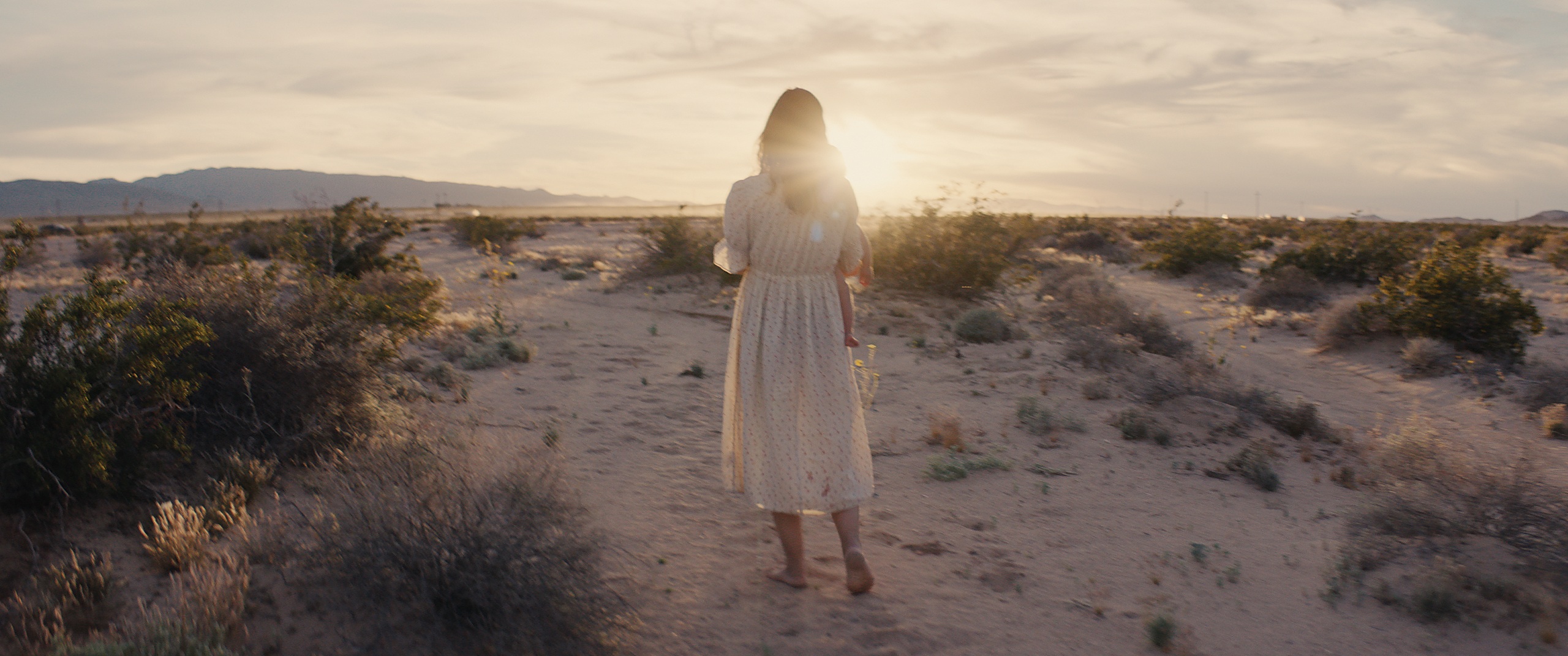 The Golden Hour — a woman in a white dress carrying a child, walking into the desert sunset, film still written and directed by Zane Ra